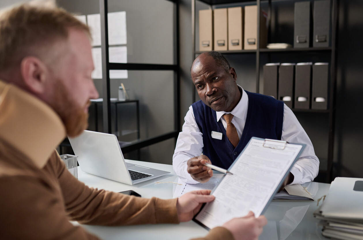 A professional advisor, who appears to be a Black man, points to a document while speaking with an injured client in a neck brace, representing the process of seeking help after an injury with no health insurance.