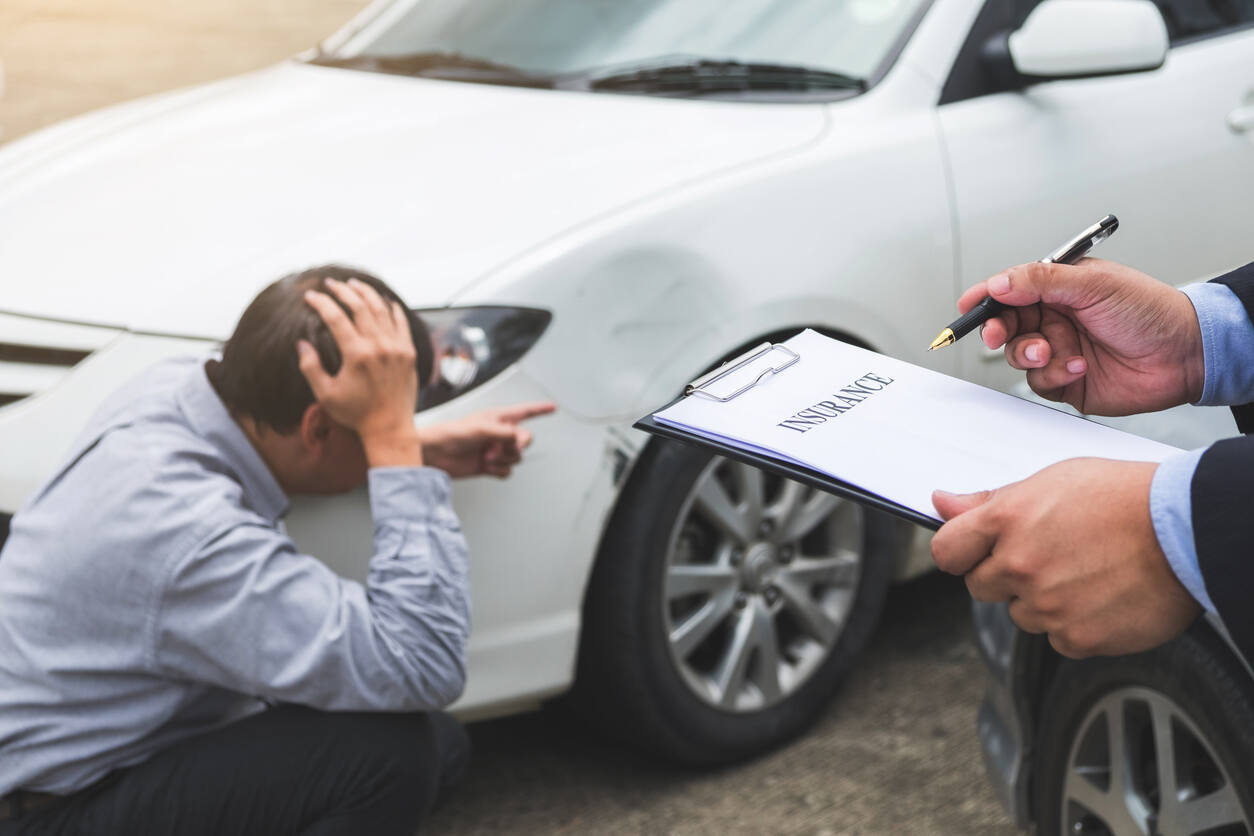 An insurance adjuster holds a clipboard while a stressed man inspects the damage to his car, illustrating the difficult decision of whether to talk to an insurance company after a Colorado accident.