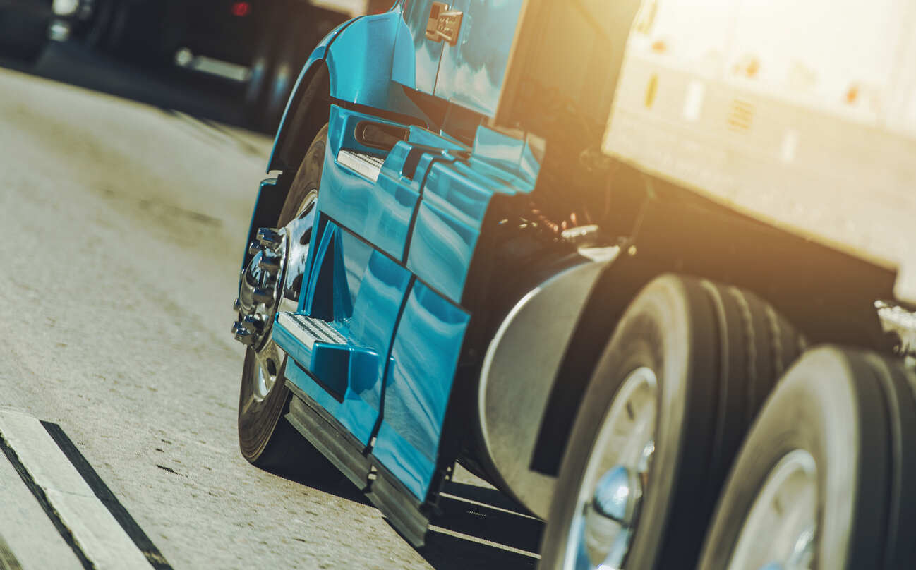 A close-up, low-angle shot of a blue semi-truck driving on a sunny street, focusing on the wheels and side panels of the truck