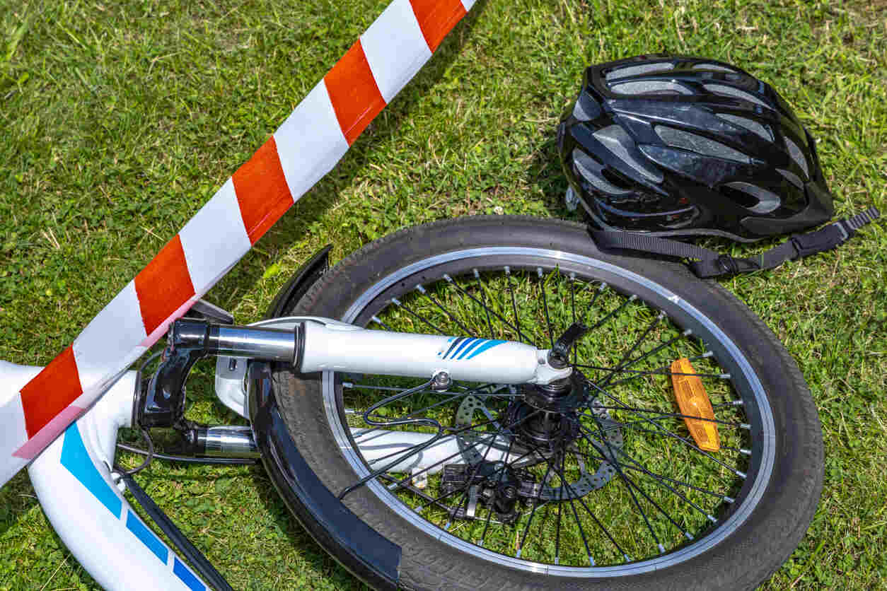 A damaged electric bicycle lying on green grass next to a black helmet and red and white caution tape, representing an e-bike accident in Denver.