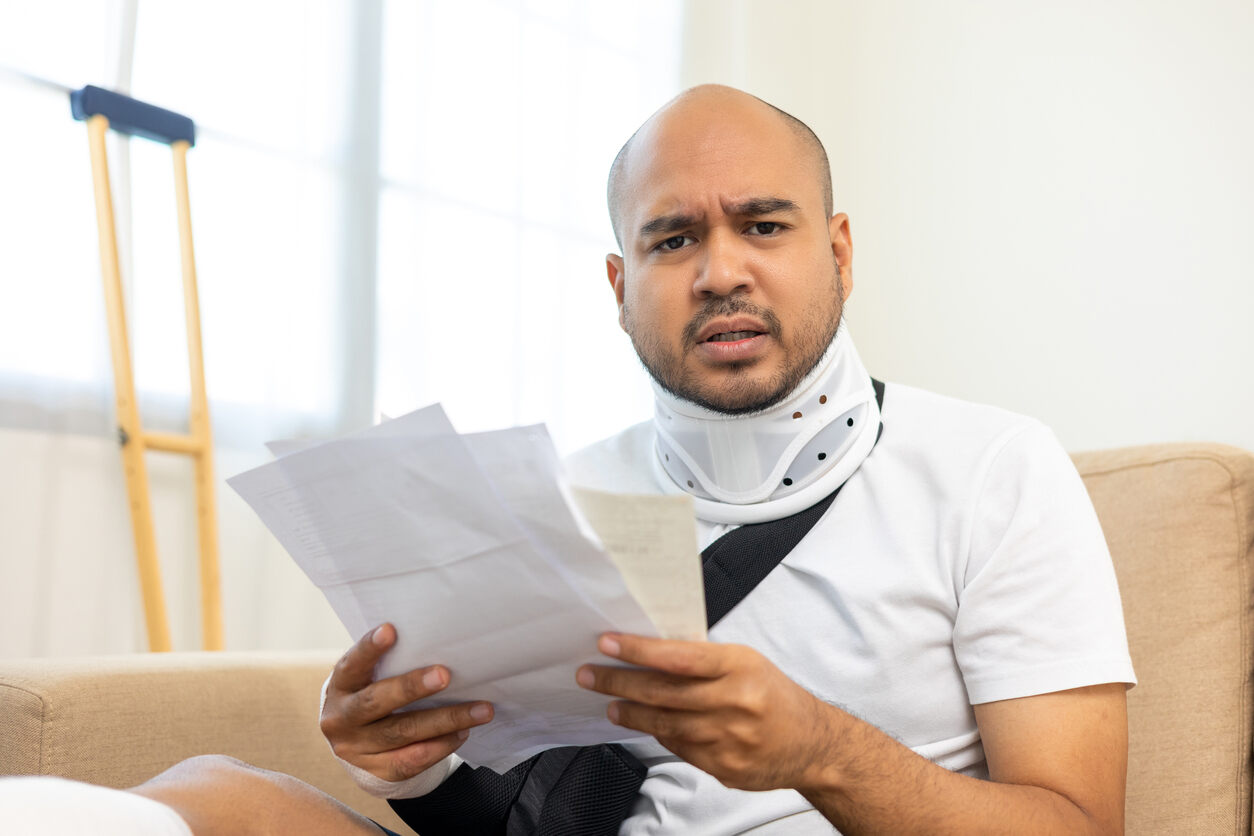 A man in a neck brace and arm sling looking at papers with a concerned expression, symbolizing lost income due to a personal injury.