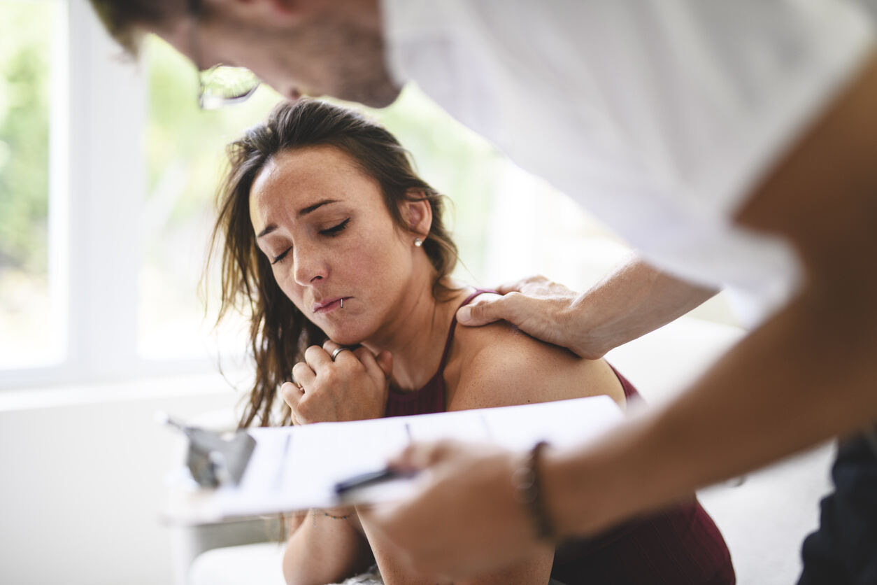 A distraught woman with furrowed brows and her hands clasped is being comforted by a person standing behind her, who has a hand on her shoulder and is holding a clipboard and pen, suggesting a difficult conversation or reporting related to potential nursing home abuse.