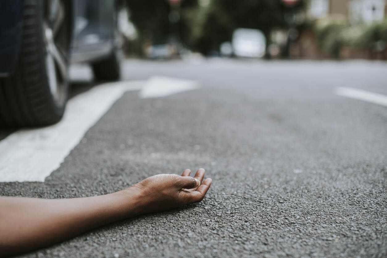 A person's outstretched hand lies on the asphalt of a street next to a car tire, implying a pedestrian accident. Road markings and a blurry background suggest a street scene in Colorado Springs.