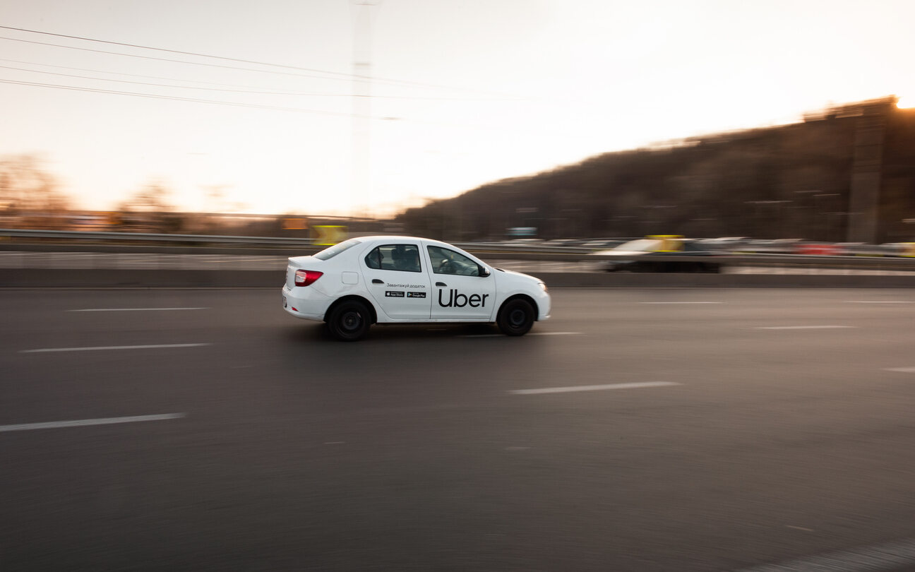 A white Uber car drives on a highway in Colorado Springs at sunset. Other blurred cars are visible in the background. The "Uber" logo is clearly displayed on the car's side.
